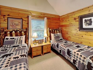 Una habitación tipo cabaña con dos camas, paredes de madera, una ventana con cortinas y una colcha con motivos de oso.