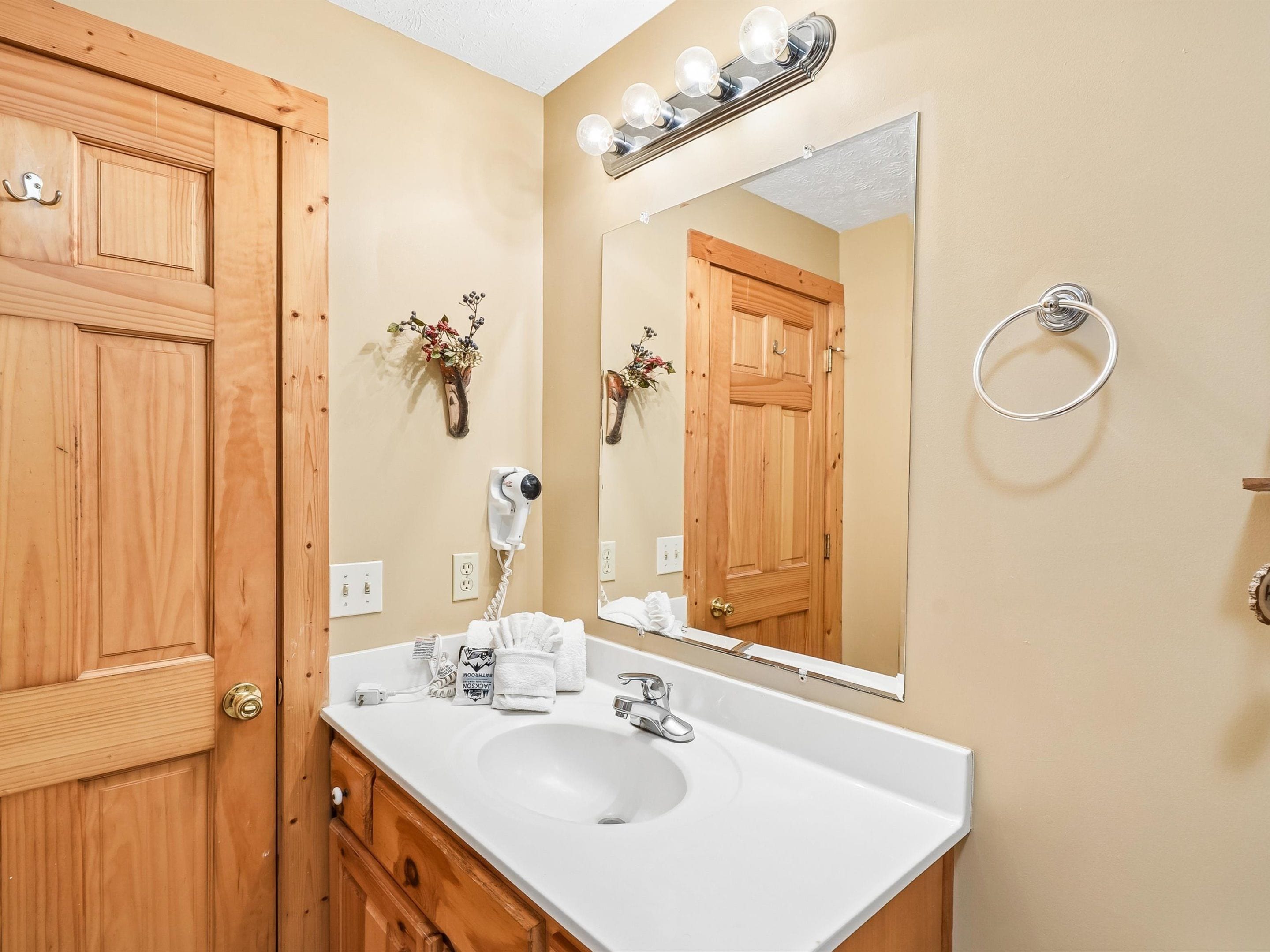 A bathroom with a wooden door, a white sink, a mirror with lights, a hairdryer, and a towel rack.