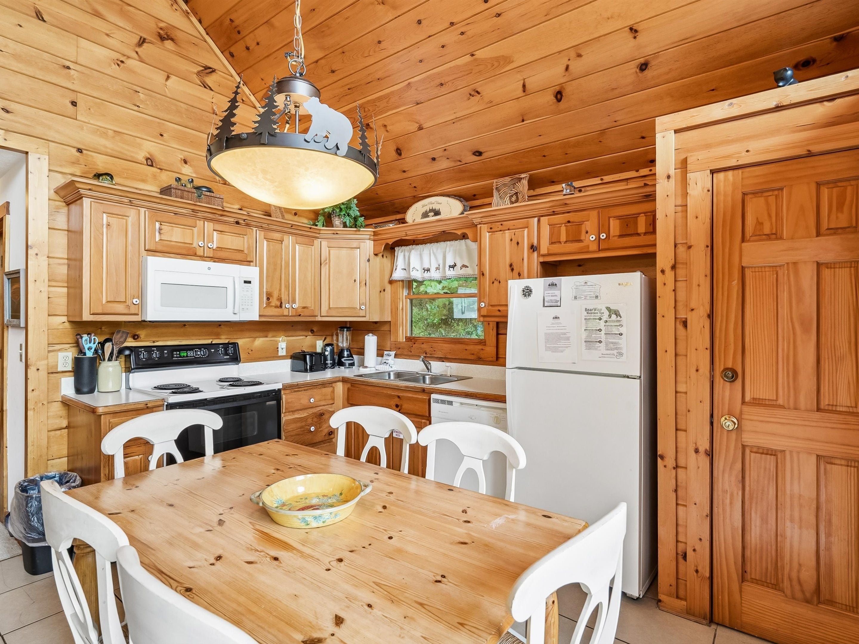 A wooden cabin kitchen with a dining table, white chairs, a stove, microwave, refrigerator, and a hanging light fixture.