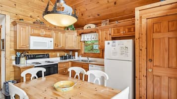 A wooden cabin kitchen with a dining table, white chairs, a stove, microwave, refrigerator, and a hanging light fixture.