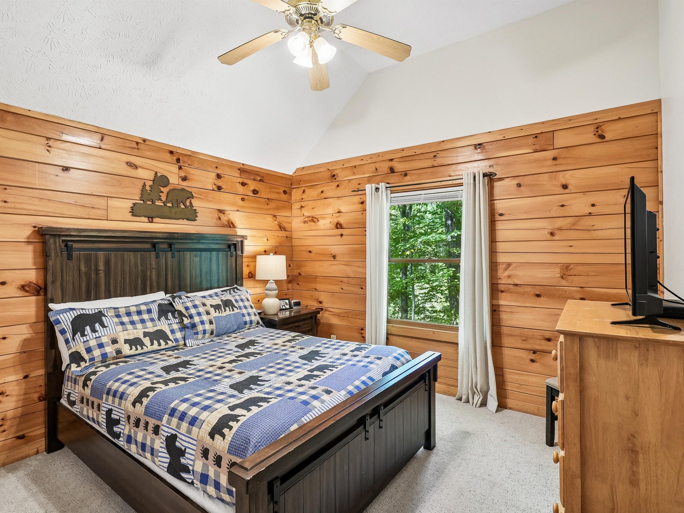 Een slaapkamer met een houten hoofdeinde, een bed met een geruit plaid, een nachtkastje met een lamp, een raam met gordijnen, en een televisie op een houten dressoir.