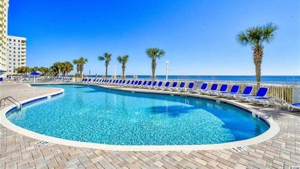 A pool with blue water, surrounded by lounge chairs and palm trees, with a clear sky and a building in the background. - NO OWNER FEES! Gorgeous Updated OCEANFRONT 2/2 Suite, OF Master, W/D, 18th Fl (North Myrtle Beach)