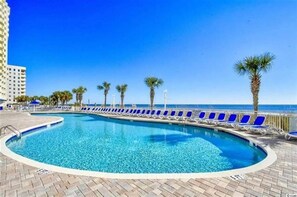 Une piscine à l’eau bleue, entourée de chaises longues et de palmiers, avec un ciel dégagé et un bâtiment en arrière-plan.