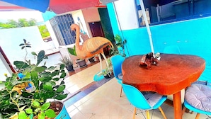 Un patio al aire libre con una mesa redonda de madera y sillas azules, rodeado de plantas en macetas y una estatua decorativa de flamenco.