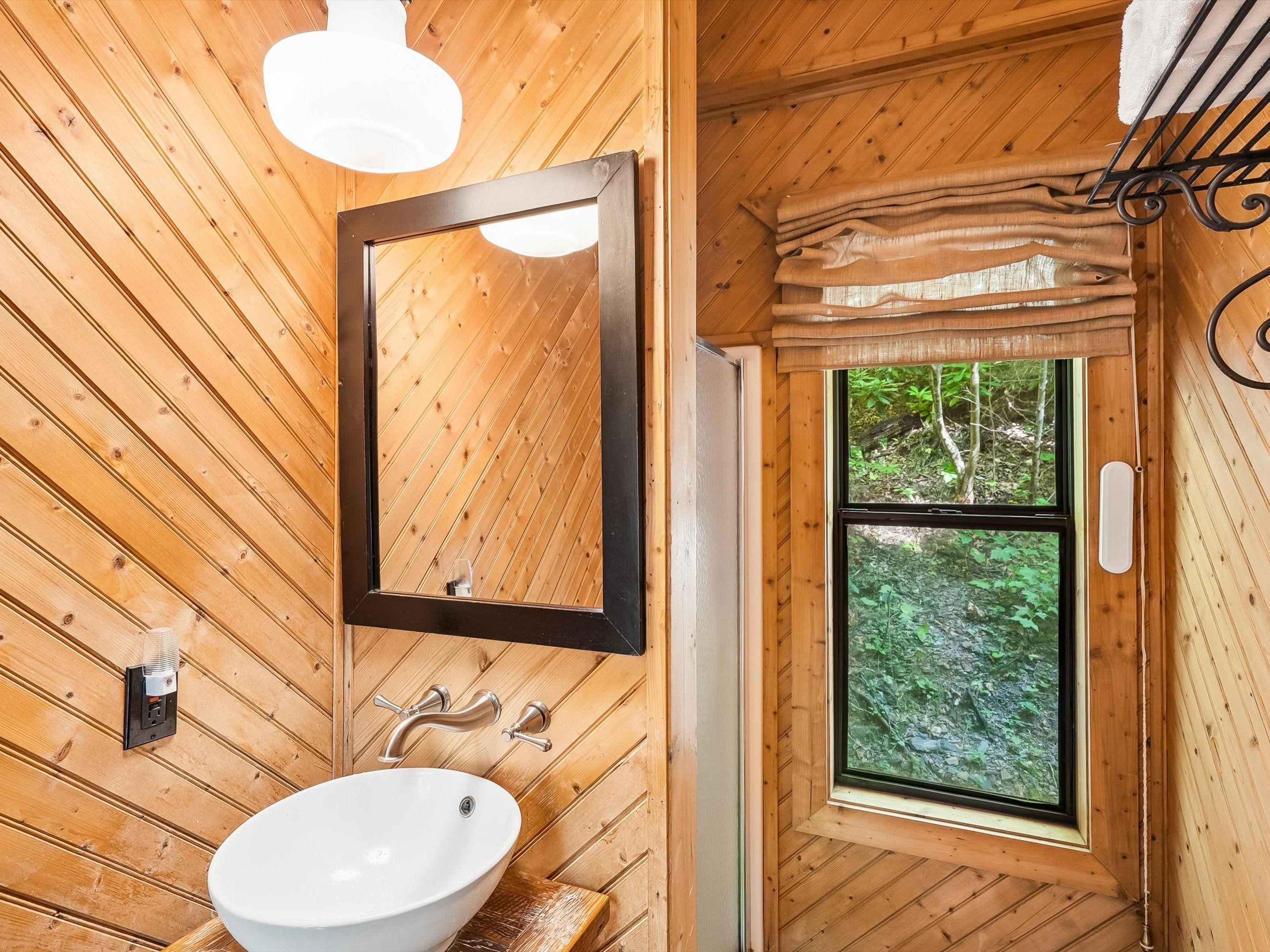 Interior de madera con un lavamanos blanco, un espejo y una ventana con vistas a la vegetación.
