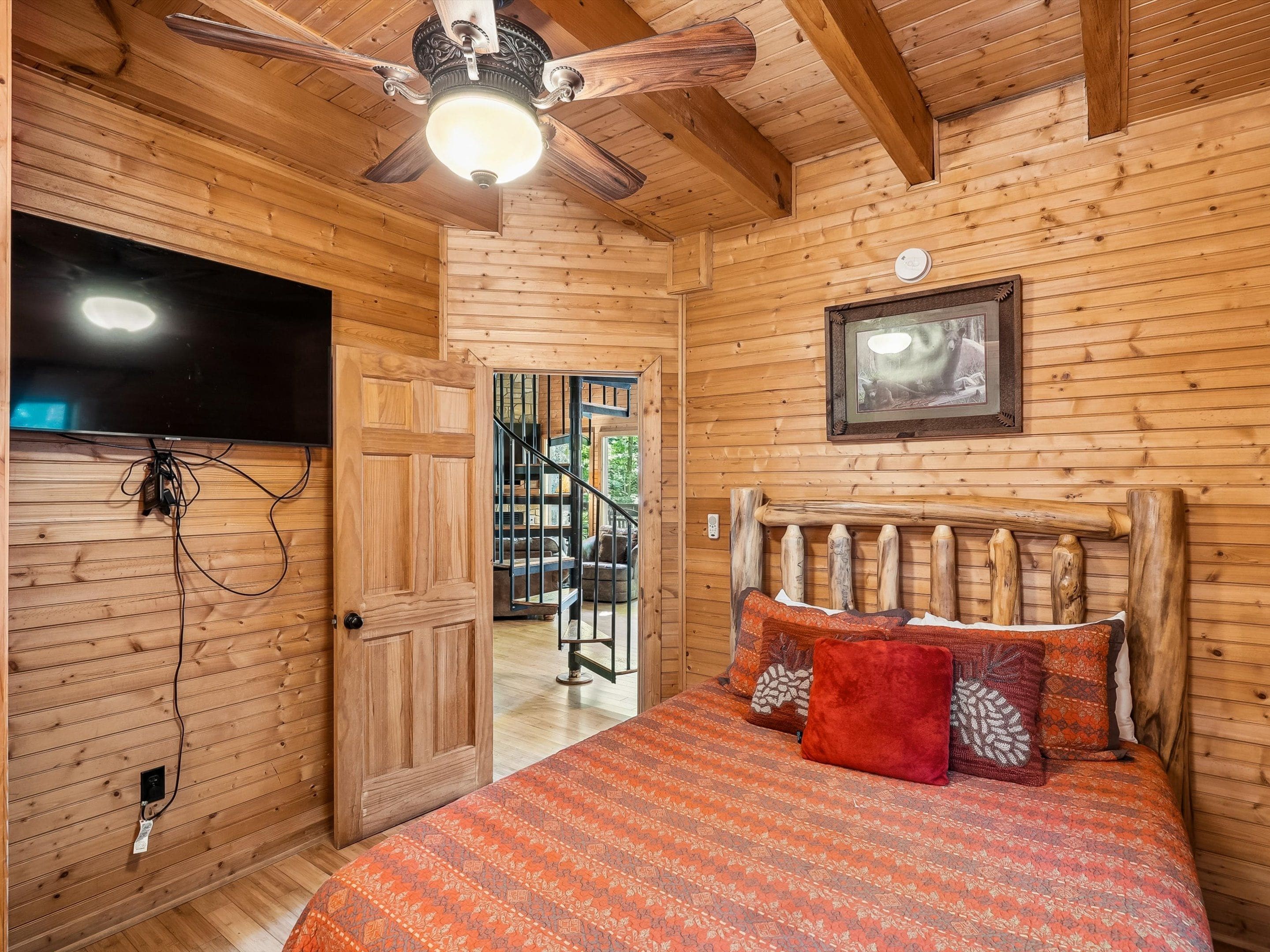 Ein Schlafzimmer einer Holzhütte mit einem Bett, einem Deckenventilator, einem montierten Fernseher und einer Treppe, die zu einem Außenbereich führt.