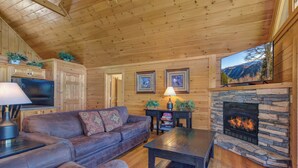 A cozy cabin interior with a stone fireplace, wooden walls, a leather sofa, and a television mounted above the fireplace. - Eagles Point Lodge by Jackson Mountain Rentals (Gatlinburg)