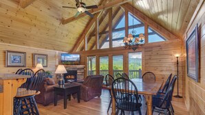 A spacious cabin interior with a high ceiling, wooden walls, a fireplace, and large windows offering a view of the outdoors. - Eagles Point Lodge by Jackson Mountain Rentals (Gatlinburg)