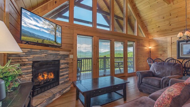 A cozy cabin interior with a stone fireplace, wooden ceiling, and large windows overlooking a deck. - Eagles Point Lodge by Jackson Mountain Rentals (Gatlinburg)
