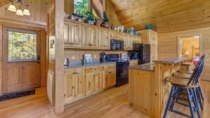 A wooden cabin kitchen with a high ceiling, wooden cabinets, a microwave, a refrigerator, and a bar area with stools. - Eagles Point Lodge by Jackson Mountain Rentals (Gatlinburg)
