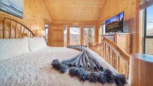 A wooden cabin bedroom with a large bed, a television mounted on the wall, and a view of the outdoors through the windows. - Eden's View by Jackson Mountain Rentals (Gatlinburg)