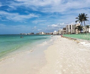 A beach with clear turquoise water, a sandy shore, and a cityscape in the background.