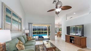 A spacious living room with a green sofa, a glass coffee table, a TV on a wooden cabinet, and a ceiling fan. - Marco Beach Haven 2 blocks from South Beach! (Marco Island)