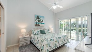 A bedroom with a bed, two bedside tables with lamps, a ceiling fan, a painting of palm trees, and a view of a pool through large glass doors. - Marco Beach Haven 2 blocks from South Beach! (Marco Island)