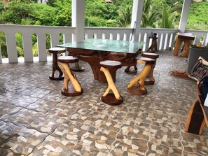 Une table en bois surmontée d’une plaque de verre, accompagnée de tabourets assortis, installée sur une terrasse carrelée, avec une clôture blanche et de la verdure en arrière-plan.