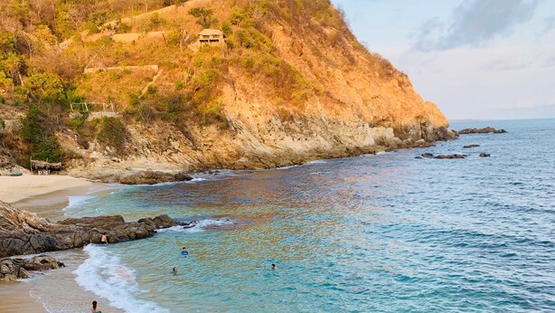 Ein Strand mit klarem türkisblauem Wasser, felsigem Ufer und einem Hügel mit spärlicher Vegetation.