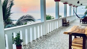 Ein Balkon mit Blick auf den Ozean bei Sonnenuntergang, weiße Säulen, eine Hängematte und ein Holztisch.