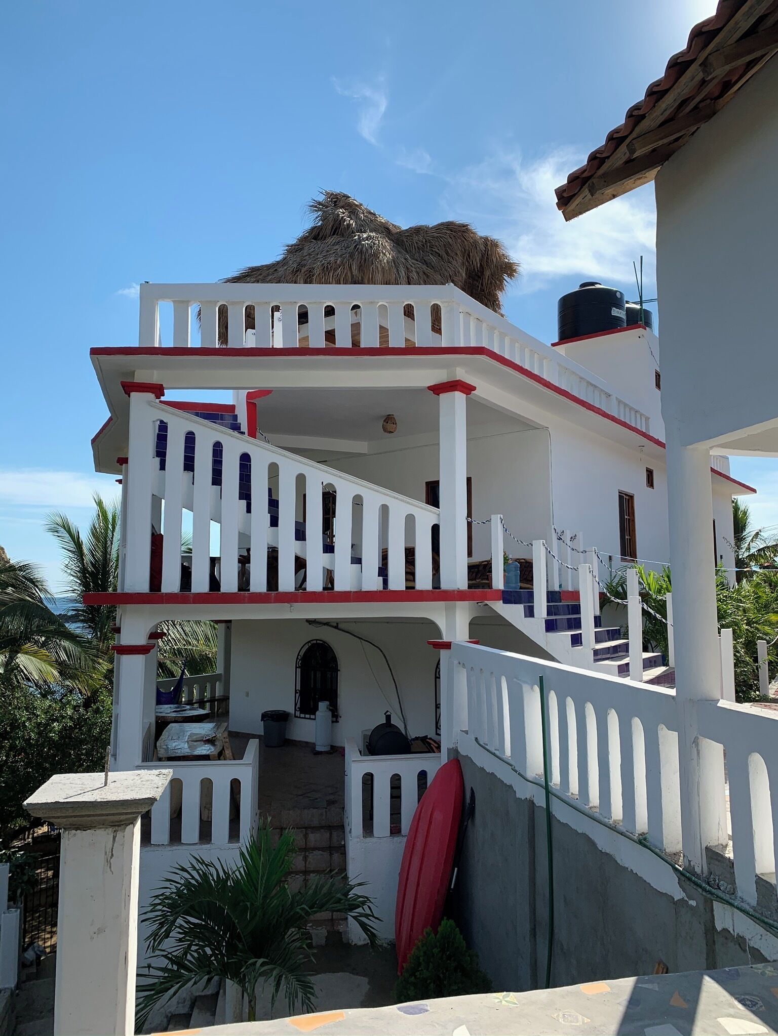 A multi-story building with a white exterior and red accents, featuring a staircase and a red surfboard leaning against the wall.