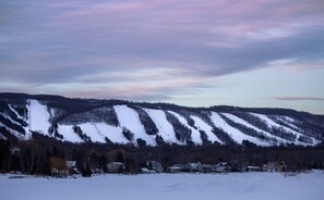 A snowy landscape with a ski slope, forested hills, and a few buildings at the base.