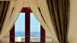 A bed with a view of the sea, curtains, a bedside table, and a lamp. - Agistri Hotel (Agistri)
