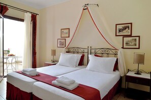 Un lit double avec du linge de lit blanc et des touches de rouge, une table de chevet avec une lampe, et une fenêtre avec des rideaux.