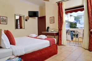 A hotel room with a bed, a desk, a chair, a TV, and a balcony with a view of buildings and greenery.