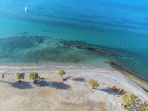 Vue aérienne d’une plage aux eaux turquoise et limpides, bordée d’une côte rocheuse et d’une végétation clairsemée.