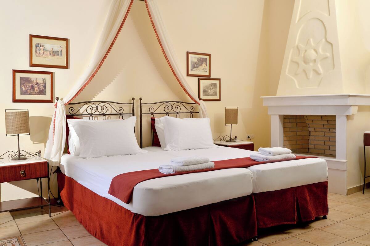 A neatly made bed with a canopy, two bedside tables with lamps, framed pictures on the wall, and a fireplace.