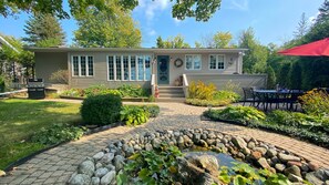 A two-story house with a front porch, a small garden with a pond, and a patio area with outdoor furniture.