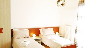Une chambre d’hôtel avec deux lits simples, une table de chevet avec une lampe, un radiateur et une fenêtre avec des rideaux.