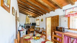 A cozy living room with a fireplace, wooden beams, a dining area with a table set for two, and a kitchen with a microwave and a tiled backsplash. - Villa Noelia Frigiliana by Ruralidays (Málaga)