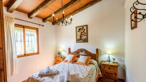 A bedroom with a wooden bed, two bedside tables with lamps, a window with curtains, and a painting on the wall. - Villa Noelia Frigiliana by Ruralidays (Málaga)