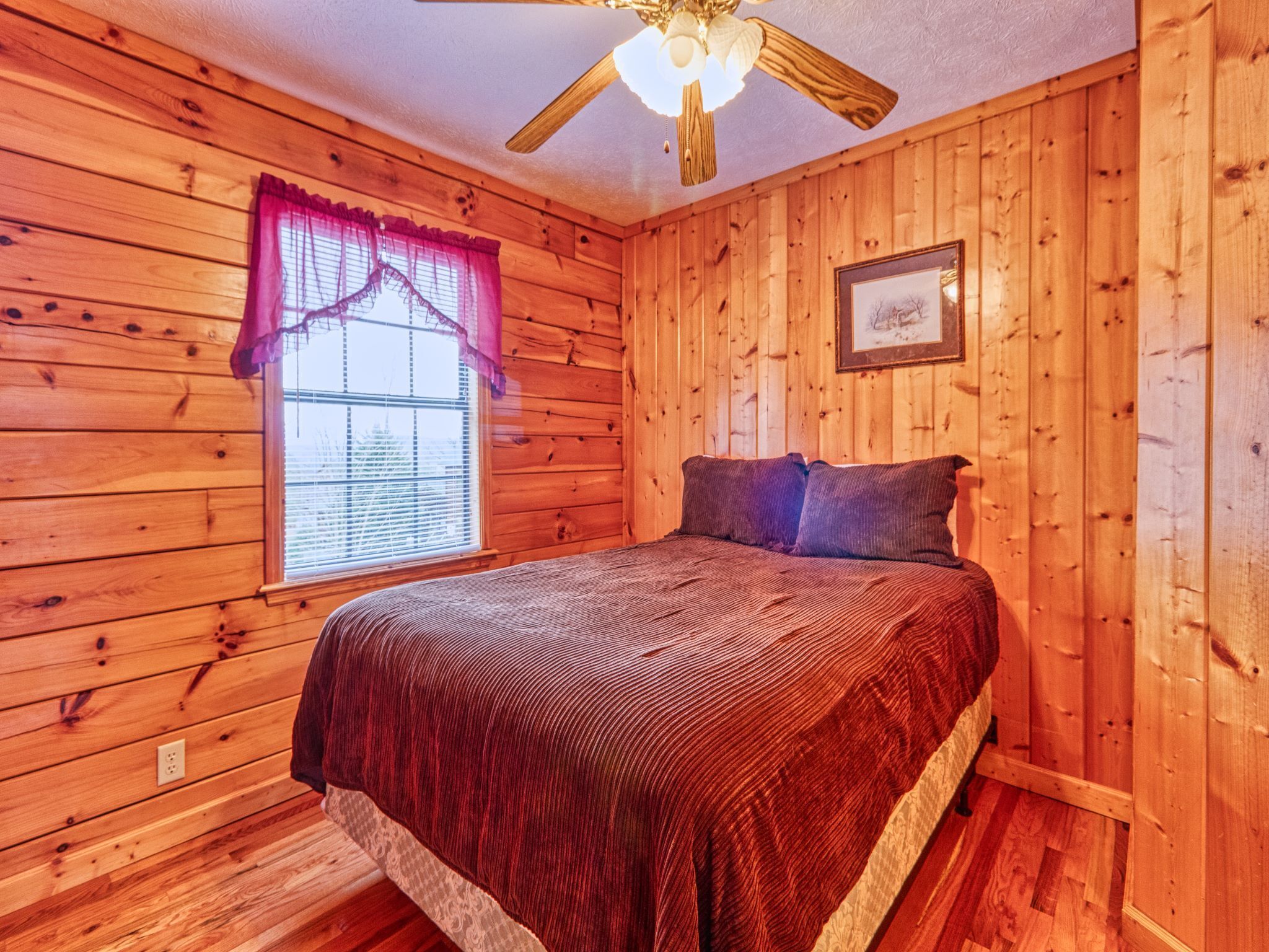 A wooden cabin bedroom with a bed, a window with curtains, a ceiling fan, and a framed picture on the wall.