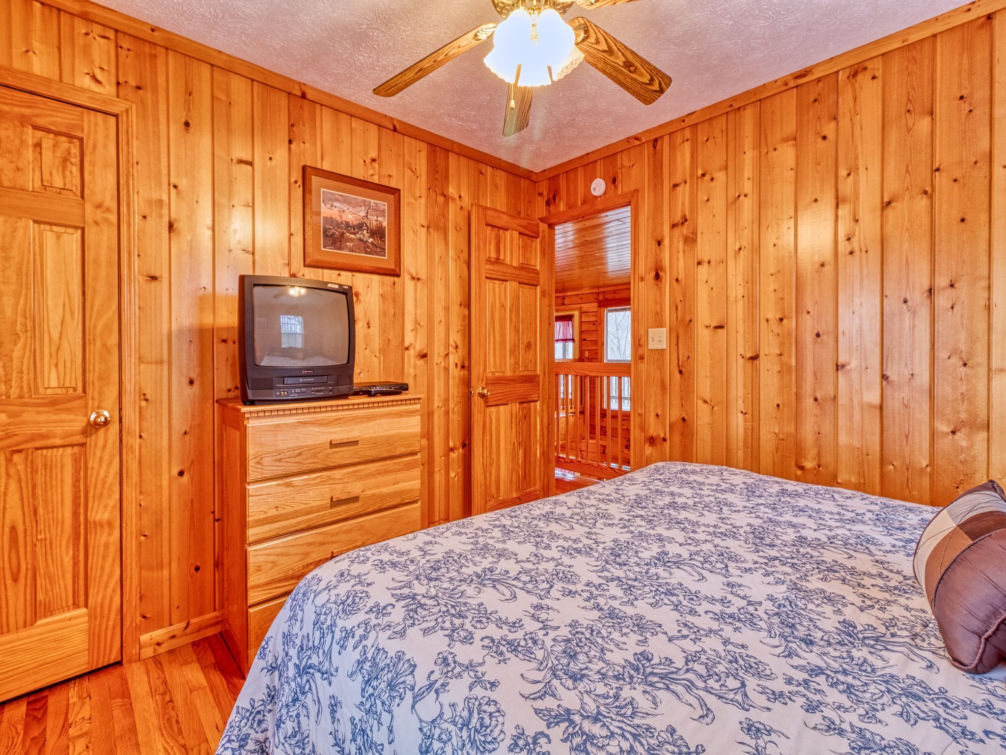 Quarto com painéis de madeira, uma cama, televisão em uma cômoda e ventilador de teto.