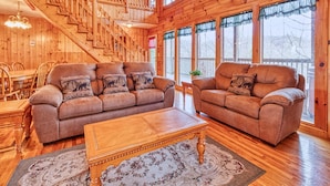 A spacious living room with a wooden staircase, two leather sofas with matching cushions, a wooden coffee table, and a large window providing a view of the outdoors. - Hawks Point Lodge by Jackson Mountain Rentals (Sevierville)