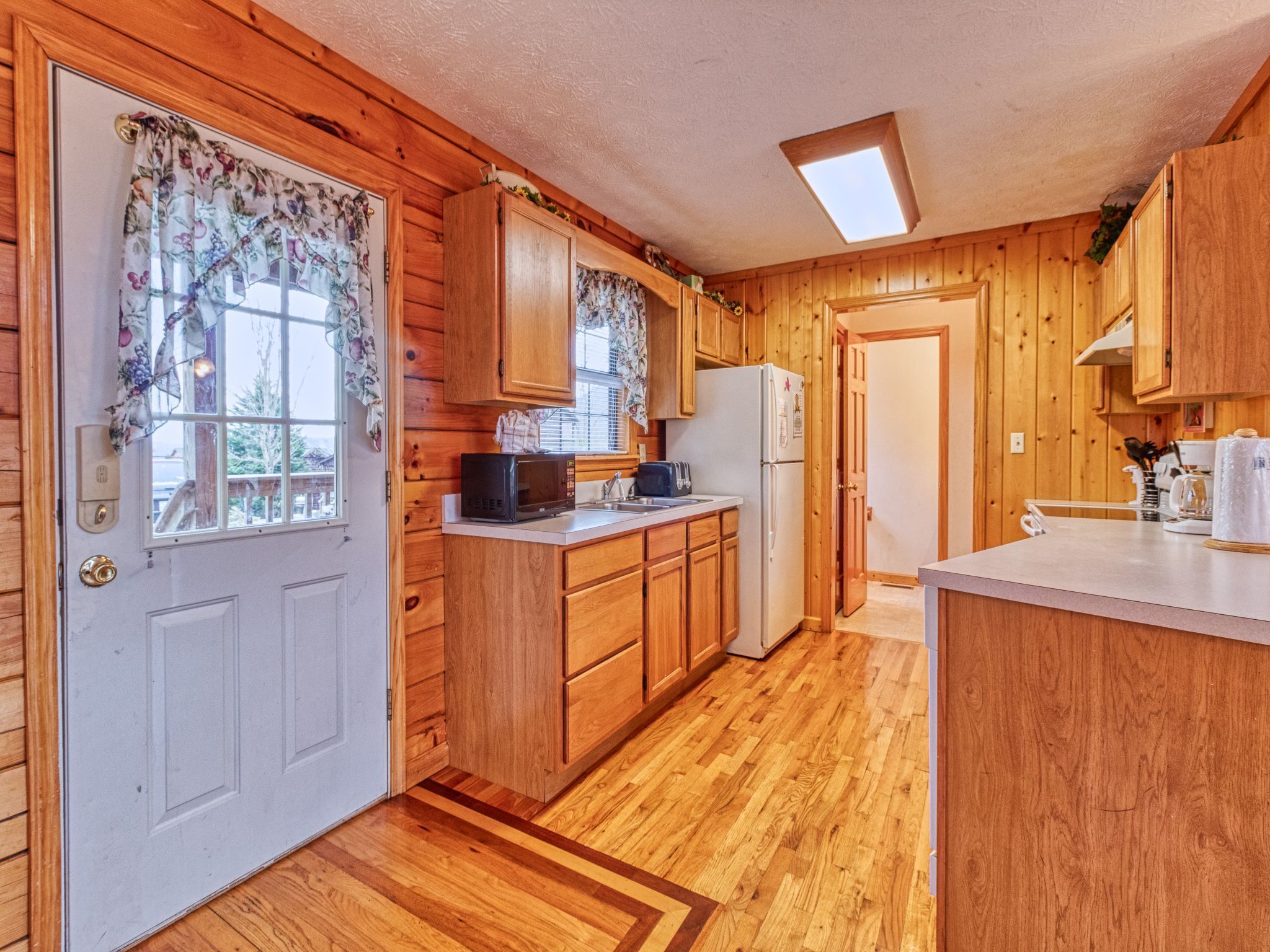 A wooden cabin-style kitchen with wooden cabinets, a microwave, a refrigerator, and a window with floral curtains.