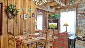 Ein rustikaler Essbereich mit Holzwänden, einem Holztisch und Stühlen. Ein Kamin, ein Fernseher und ein Fenster mit Vorhängen sind ebenfalls vorhanden.