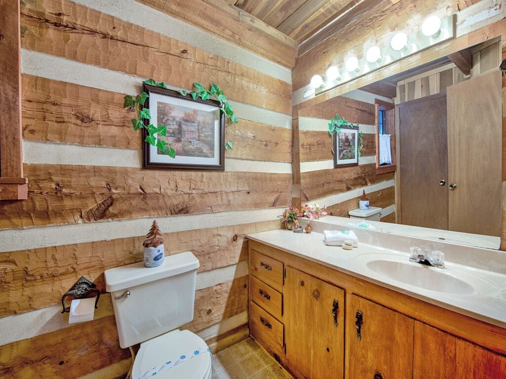A rustic bathroom with wooden walls, a white toilet, a wooden vanity with a mirror, and framed pictures on the wall.