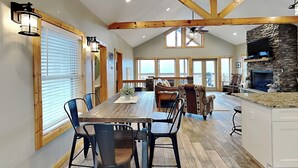 A spacious dining area with a long wooden table and chairs, a kitchen with a stone fireplace, and large windows providing natural light.