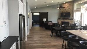 A modern kitchen with white cabinets, a large island with bar stools, a stone fireplace, and a dining area with a dark wood table and chairs. - Beautiful gated lakefront home built im 2020 (Mabank)