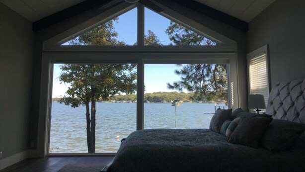 A bedroom with a large window overlooking a lake, a bed with multiple pillows, and a ceiling fan. - Beautiful gated lakefront home built im 2020 (Mabank)