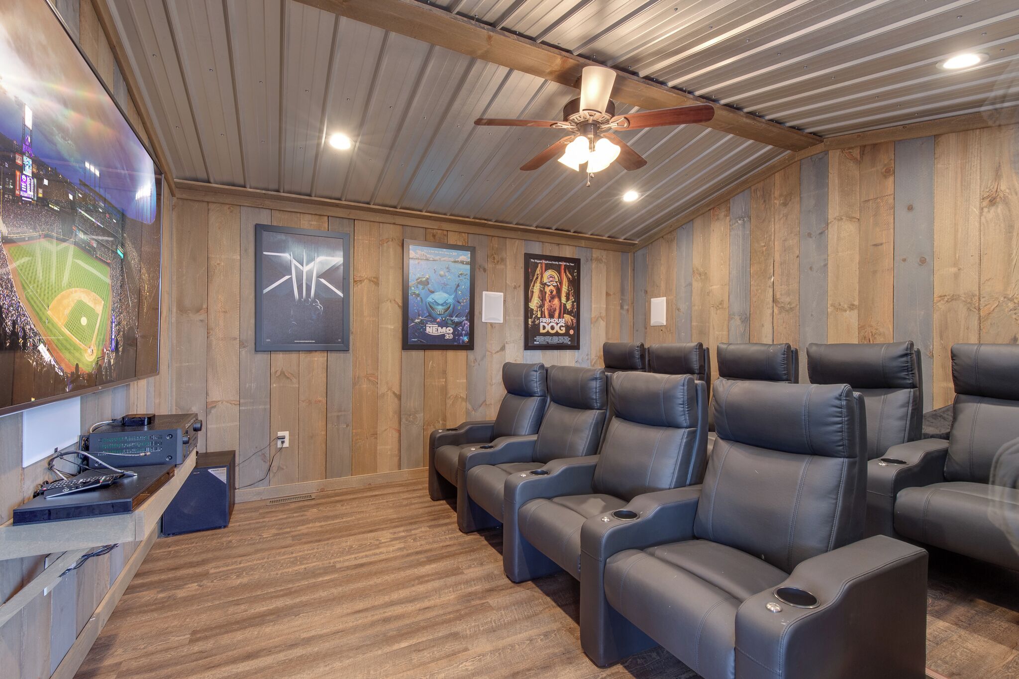 A home theater room with wooden walls, ceiling fan, reclining chairs, and a large screen displaying a baseball game.