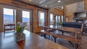 A wooden interior with a dining area, a stone fireplace, and large windows offering a view of the mountains. - Great Smoky Lodge by Jackson Mountain Rentals (Gatlinburg)