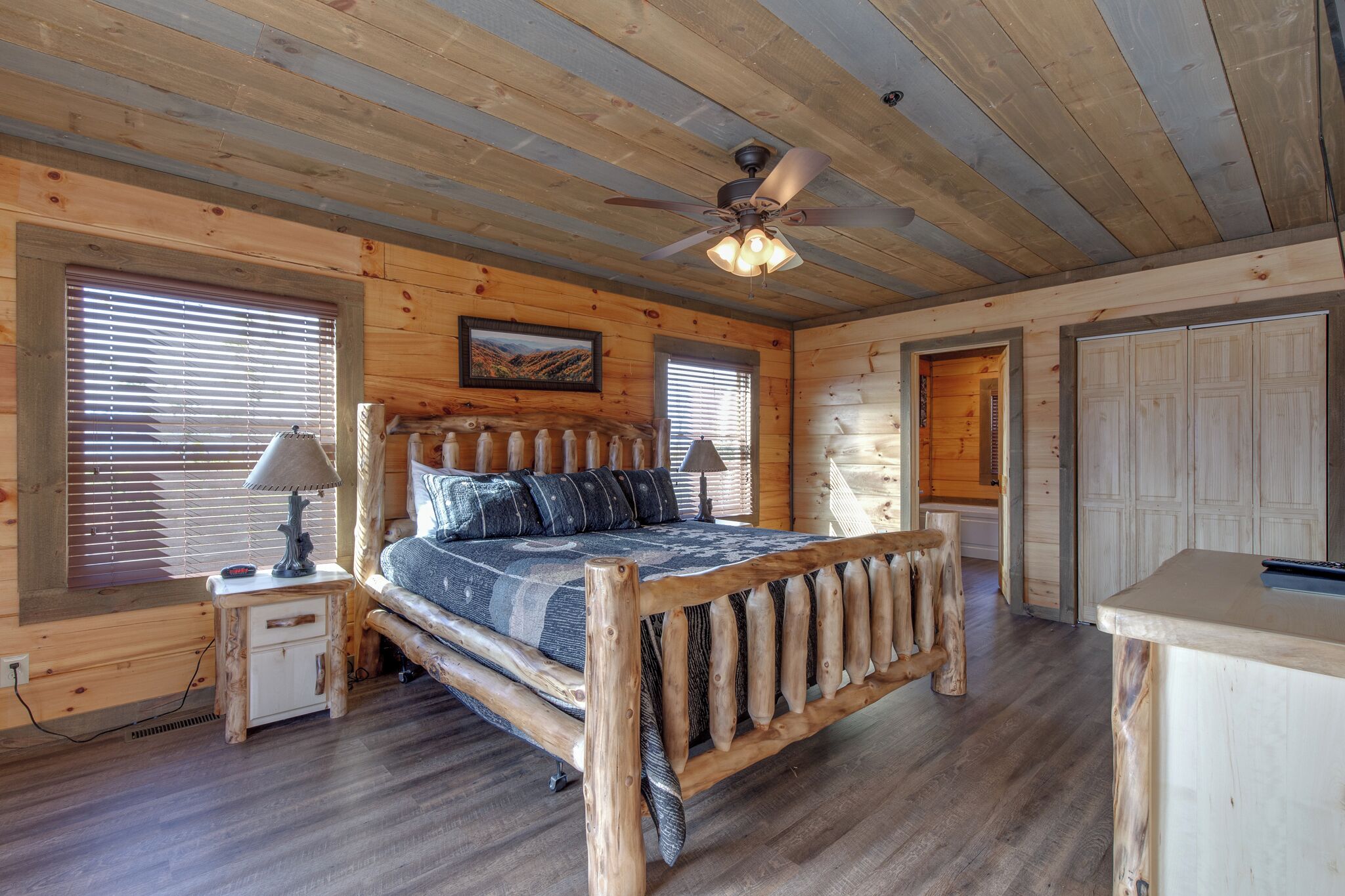 A log cabin bedroom with a wooden bed, two bedside tables, a ceiling fan, and a large window with blinds.