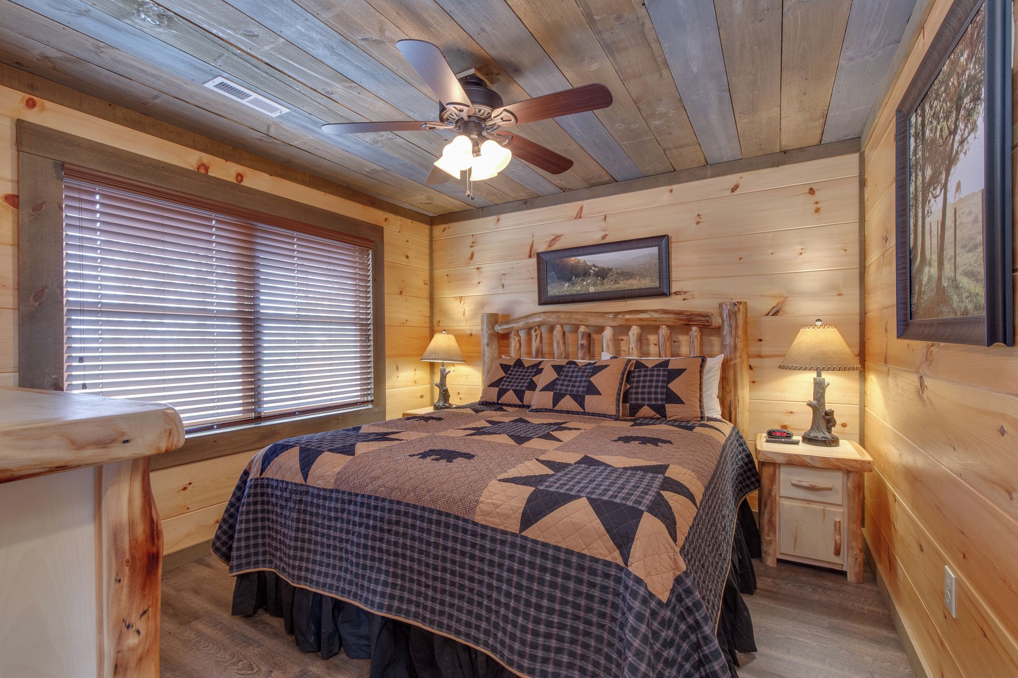 A cozy wooden cabin bedroom with a bed, two bedside tables with lamps, a ceiling fan, and a window with blinds.