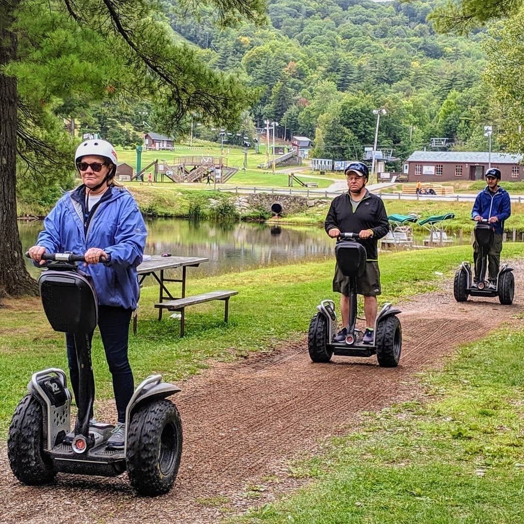 Kolme ihmistä ajaa segway-lautoilla järven lähellä. Taustalla näkyy piknikpöytiä ja leikkikenttä.