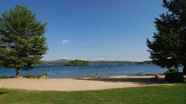 Ein See mit einem Sandstrand, einer grünen Wiese, Bäumen und freiem Blick auf das Wasser.