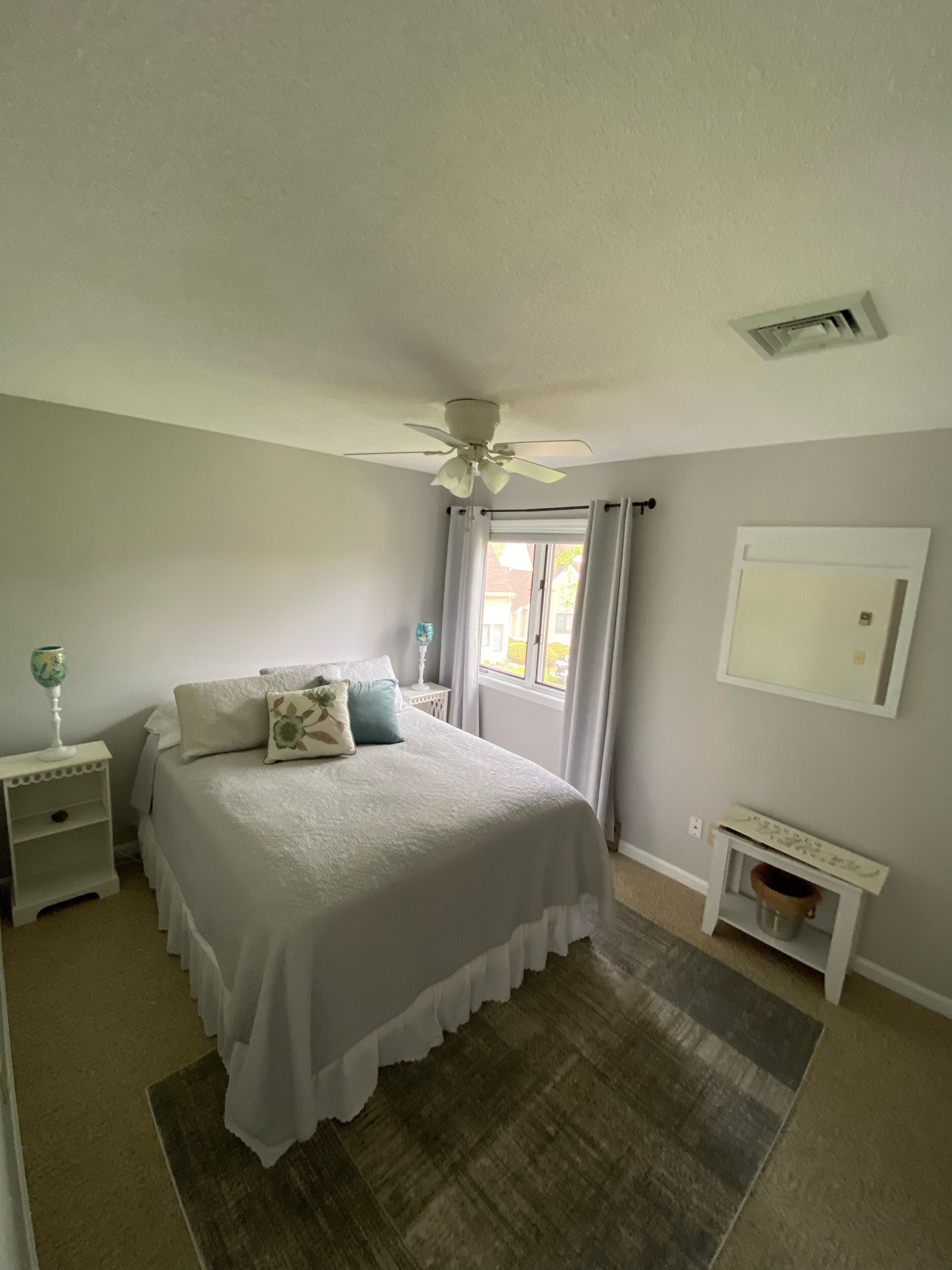 Un dormitorio con cama, mesitas de noche, ventilador de techo, ventana con cortinas, una pequeña mesa y un espejo.