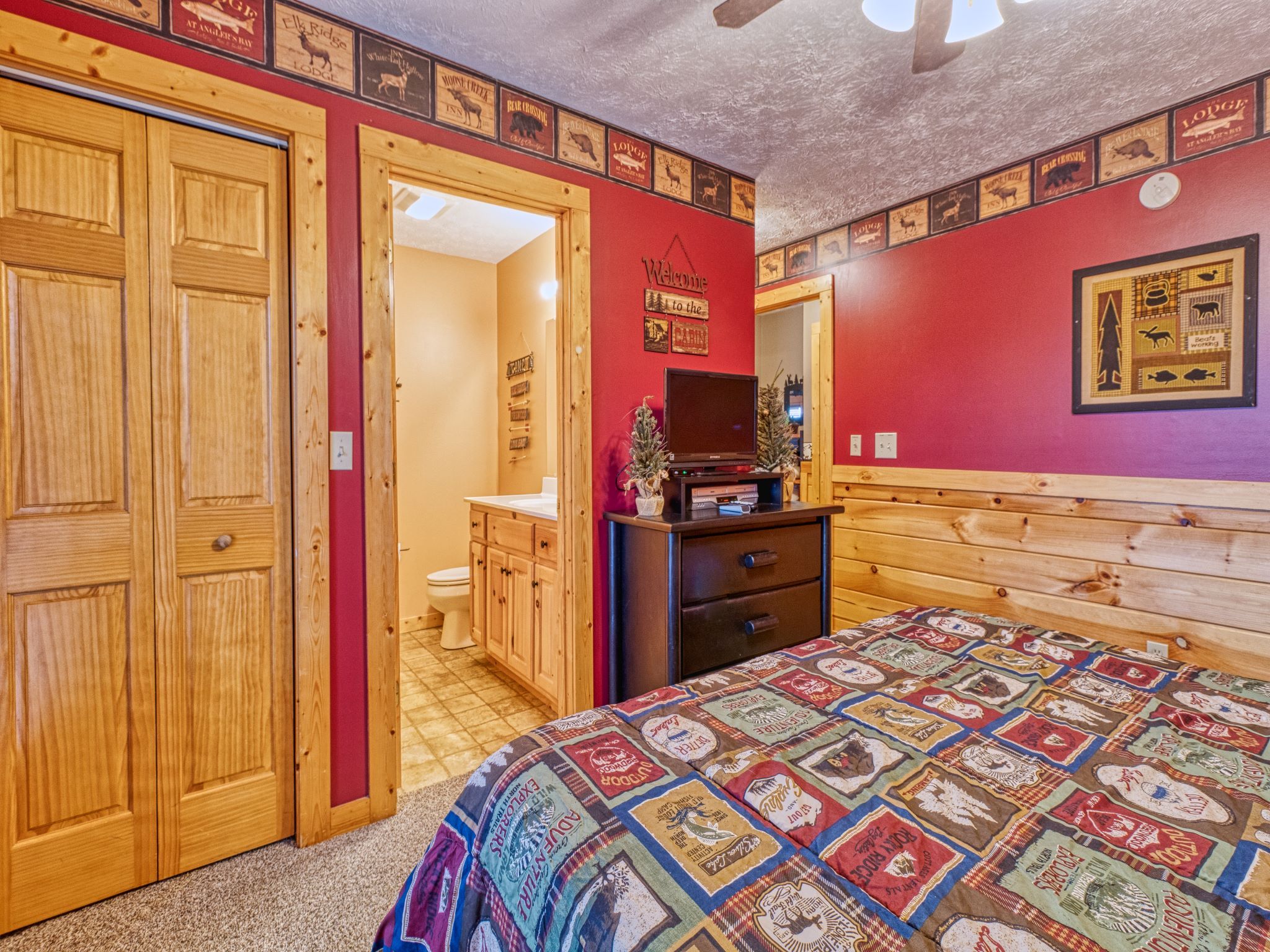 Ein Schlafzimmer mit einem hölzernen Bett, einem Schminktisch und einem Fernseher auf einem Schrank. Eine Tür führt ins Badezimmer, und es gibt einen Kleiderschrank.