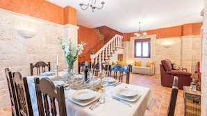 Een eetgedeelte met een tafel voor vier personen, een trap en een woonkamer met een bank en kussens.
